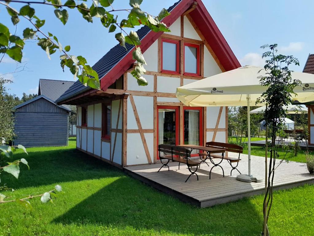 A charming wooden home with a sloped roof features prominent red window frames. A spacious deck provides seating for outdoor dining under a large umbrella. The surrounding grassy area includes trees, enhancing the natural setting.