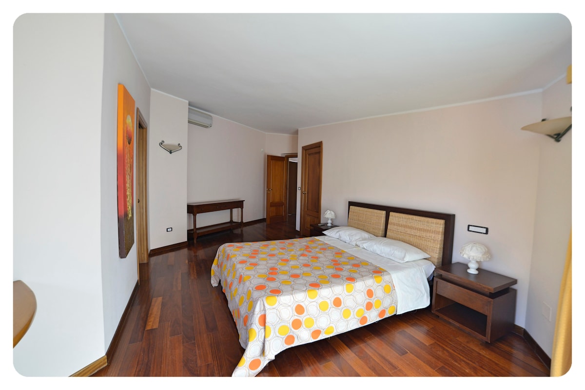 A bedroom is furnished with a spacious bed featuring a patterned blanket. Wooden flooring complements the light-colored walls, while bedside tables and lamps are positioned beside the bed. A small table is visible near the entrance, and a door leads to additional areas.