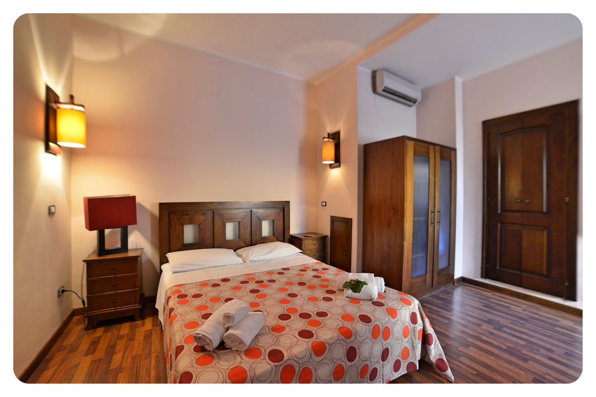 A modern bedroom is presented with a spacious bed covered in a polka dot blanket. Wooden furnishings, including nightstands and a wardrobe, provide functional storage. Soft lighting from wall sconces creates a warm atmosphere, complemented by air conditioning for comfort.