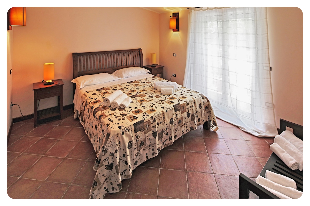 A spacious sleeping area features a comfortable bed dressed with a decorative blanket. Soft lighting is provided by lamps on either side of the bed. Natural light filters through a large window covered by sheer curtains, illuminating the warm tones of the room.