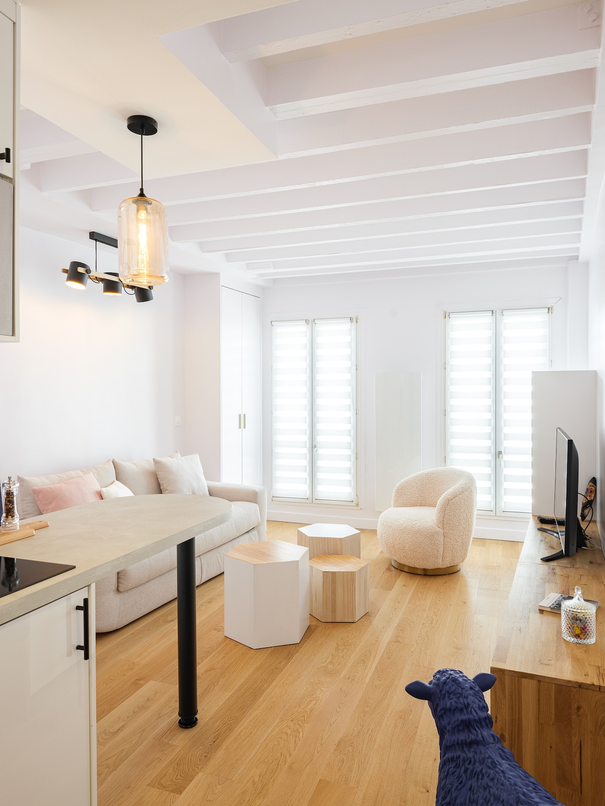 The living area features a light-colored sofa and a circular armchair, complemented by contemporary wooden furniture. Two wooden stools are placed in front of a small table. Large windows with louvered shutters allow for natural light to fill the space.