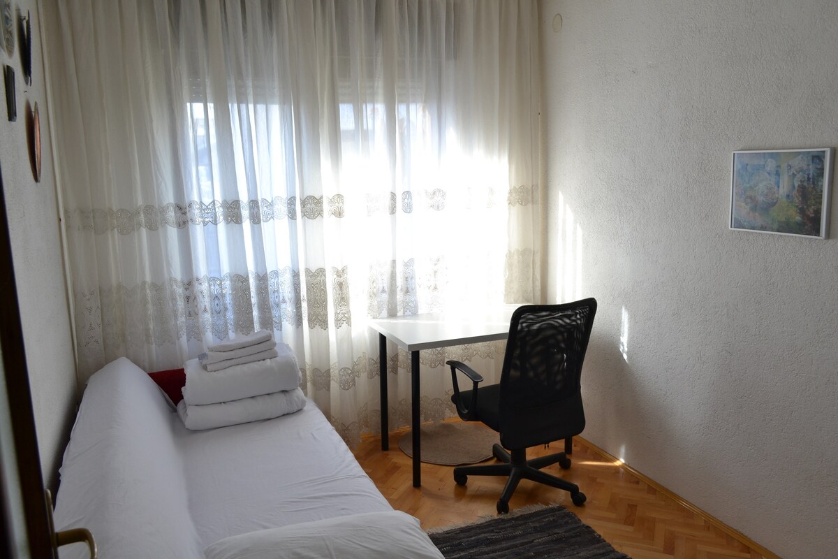 A bright bedroom features a large window draped with sheer curtains, allowing natural light to fill the space. A spacious bed is neatly arranged with folded linens, complemented by a desk and an office chair situated nearby. A framed artwork decorates the wall.