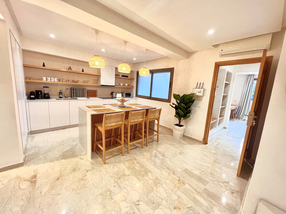 The kitchen area features modern cabinetry and a spacious island with four rattan bar stools. Natural light floods the space through a large window, highlighting the marble flooring. Appliances such as a coffee maker and microwave are neatly arranged on the countertop.