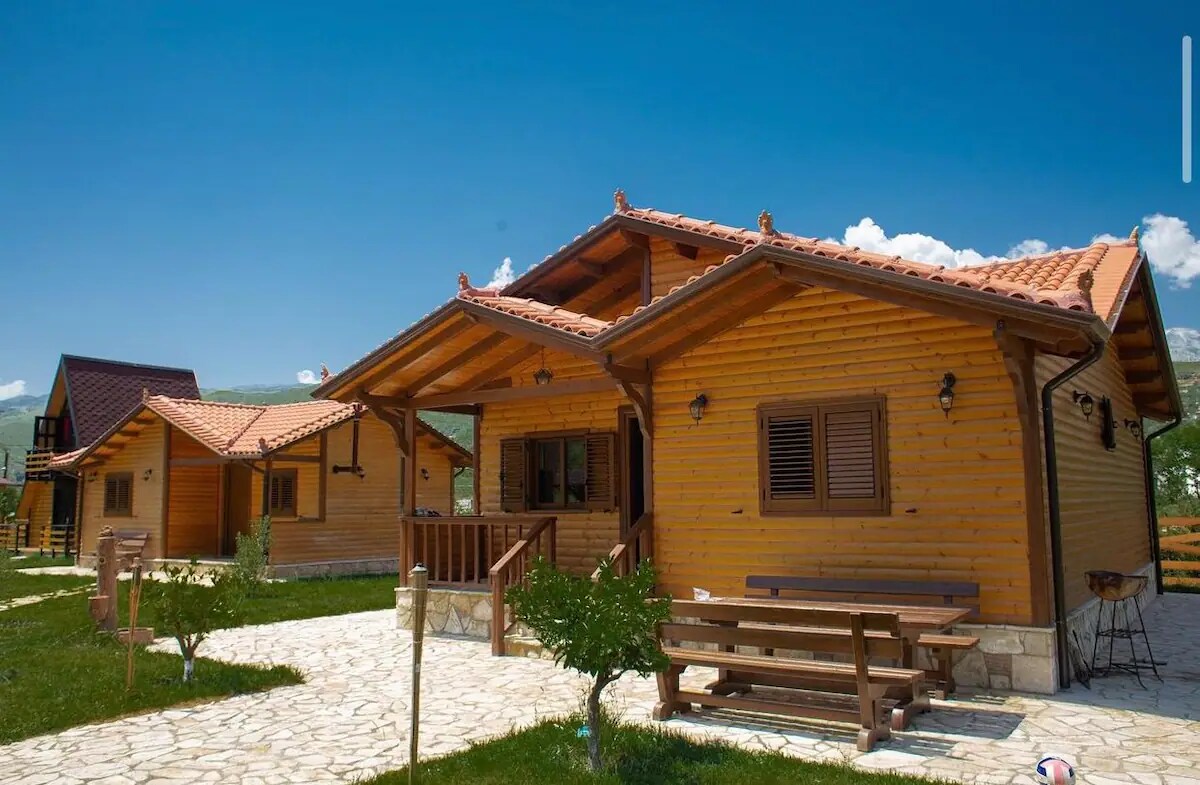 A wooden house with a warm, inviting facade features a covered porch with seating. The natural stone path leads through a grassy area, flanked by other similar structures, under a clear blue sky with subtle clouds.