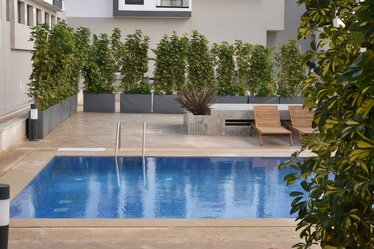A serene pool area is presented with clear blue water, surrounded by neatly trimmed greenery and planters. Two wooden lounge chairs are positioned alongside the pool, inviting relaxation in a private outdoor setting.