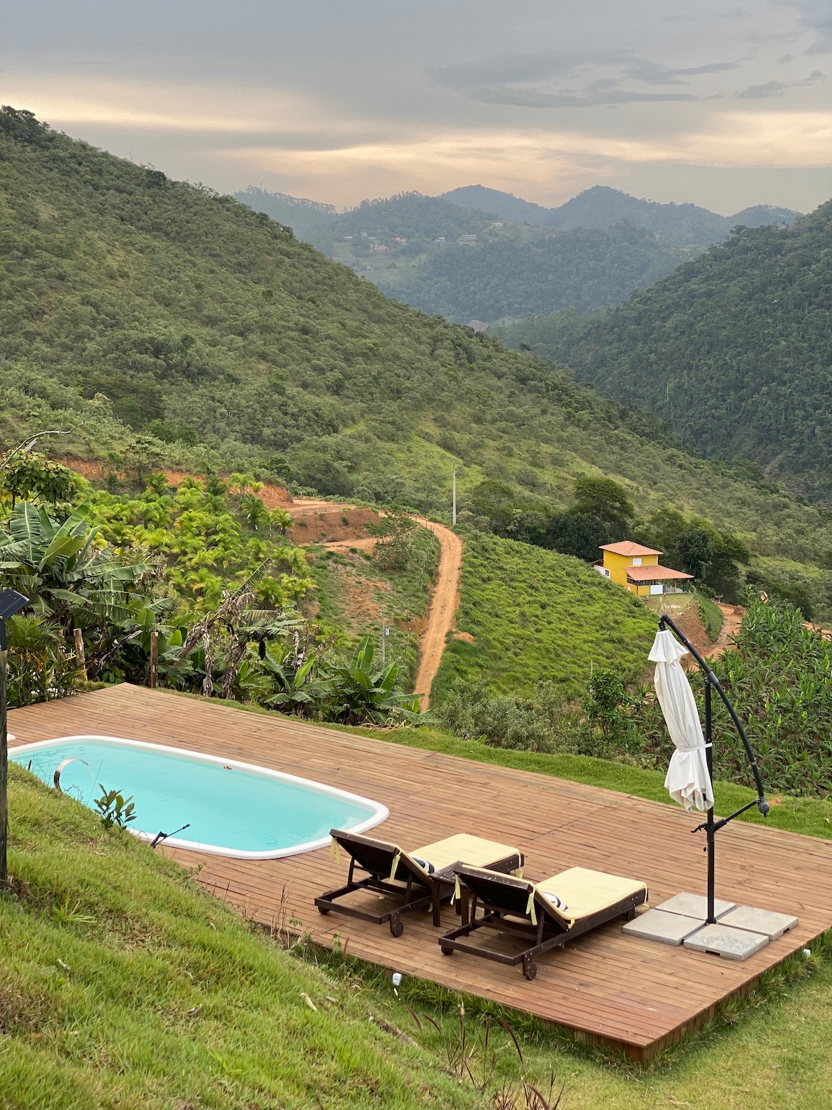 An outdoor pool is situated on a wooden deck, offering a clear view of lush green hills and mountains in the background. Two lounge chairs are placed beside the pool, and a parasol stands nearby, providing a shaded area.