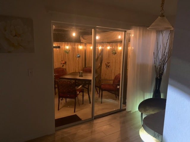 A dining area is visible through glass sliding doors, featuring a wooden table surrounded by red cushioned chairs. Soft lighting from string lights creates a warm ambiance, while bamboo accents provide a cozy outdoor feel. Natural light filters in through a nearby sheer curtain.