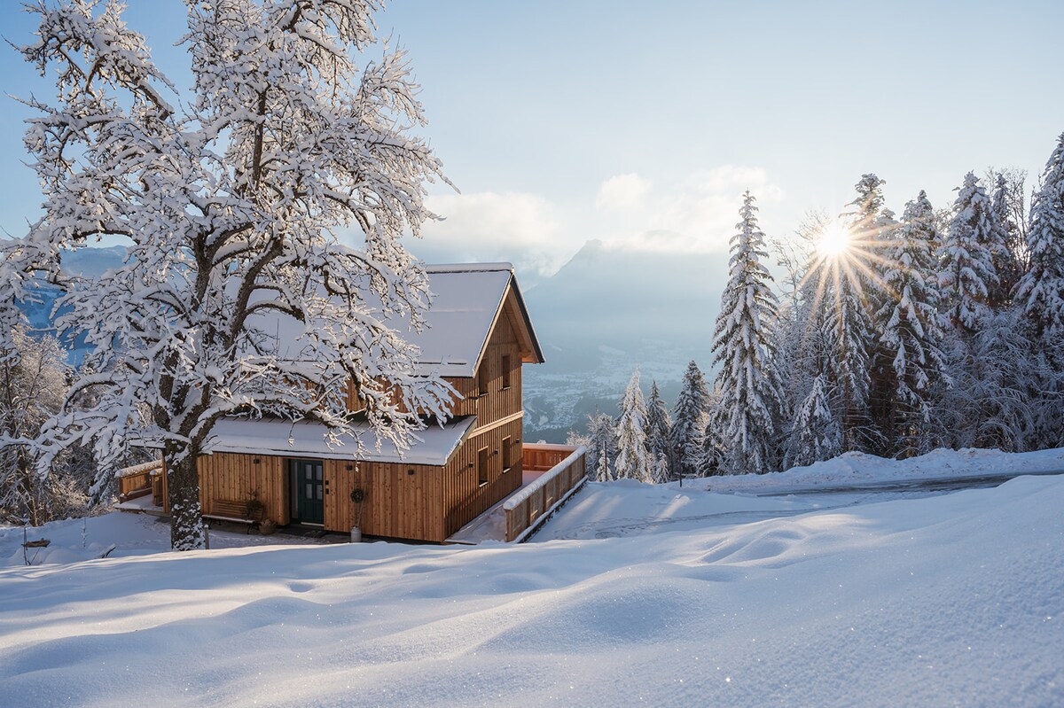 A chalet is nestled among snow-covered trees, showcasing wooden architecture. The landscape features a blanket of pristine snow, with mountains in the background. Sunlight streams through the trees, creating a serene winter atmosphere.