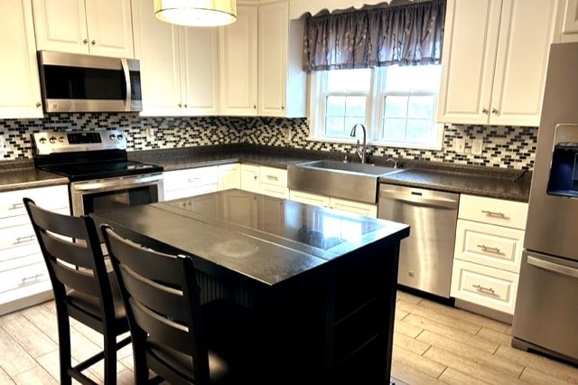 A modern kitchen is equipped with stainless steel appliances and a spacious island with seating for two. Light cabinetry contrasts with dark countertops, while a mosaic backsplash adds texture. Natural light filters through two windows, enhancing the bright and functional cooking space.