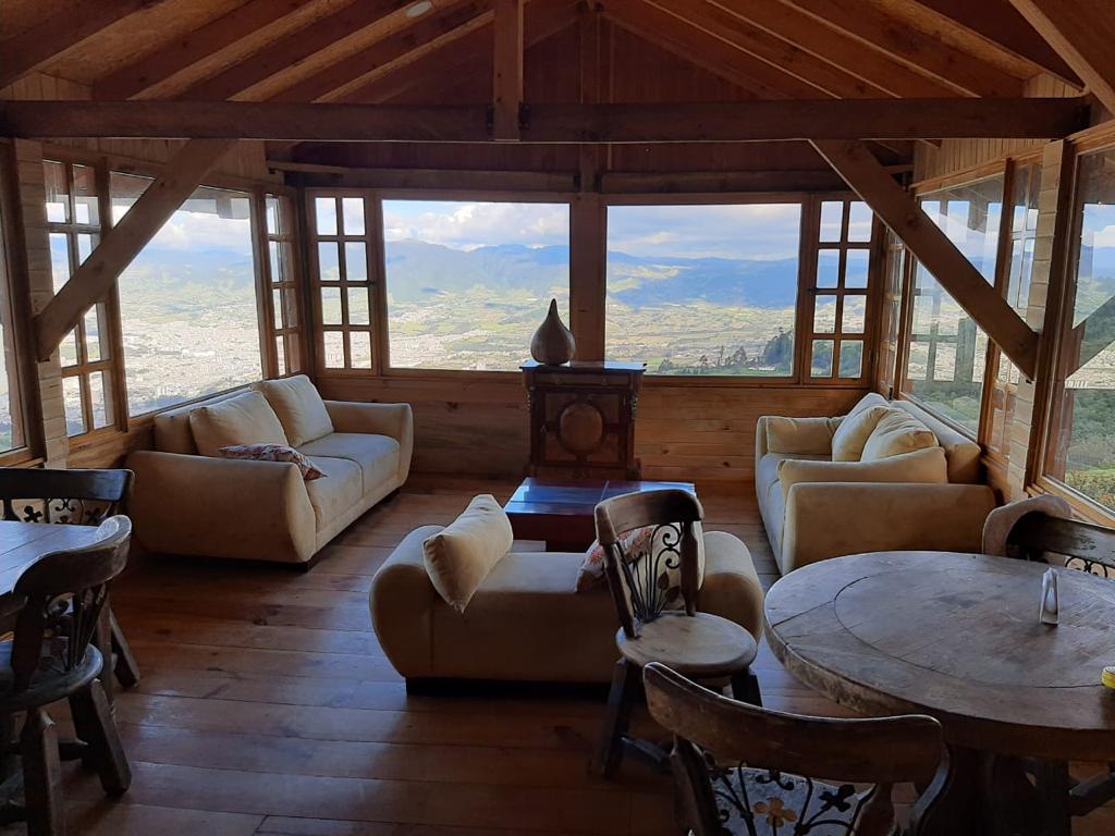 The spacious living area features large windows that provide panoramic views of the surrounding landscape. Comfortable seating is arranged thoughtfully, complemented by a wooden coffee table in the center. A rustic dining table is visible, enhancing the cabin's warm ambiance.