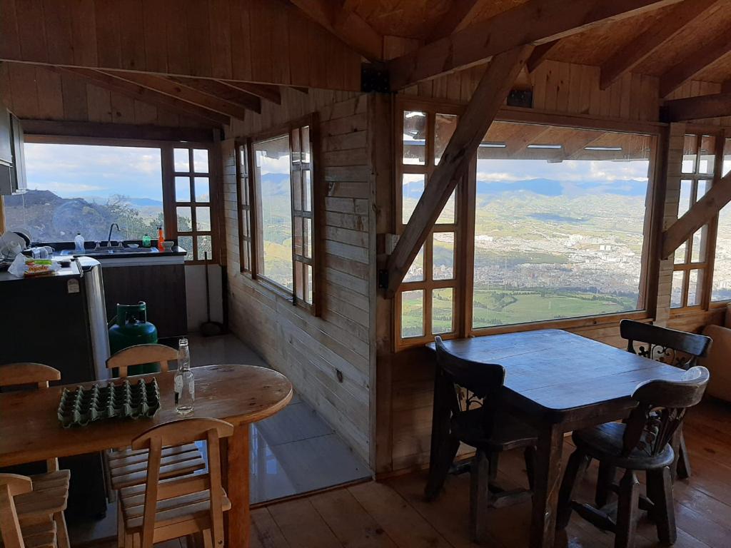 A wooden interior features large windows that provide panoramic views of the surrounding landscape. A dining table with chairs is positioned near the window, while a kitchenette is slightly visible in the background, with natural light flooding the space.