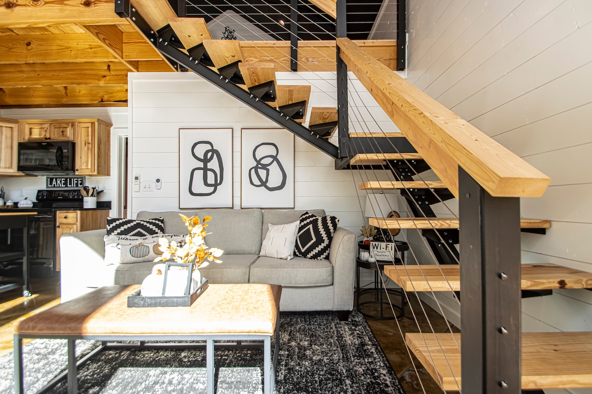 An open living space features a modern staircase with wooden steps and metal railing. A light gray sofa is positioned near a coffee table adorned with decorative items. Two abstract wall art pieces are displayed above the sofa, while warm sunlight illuminates the room.