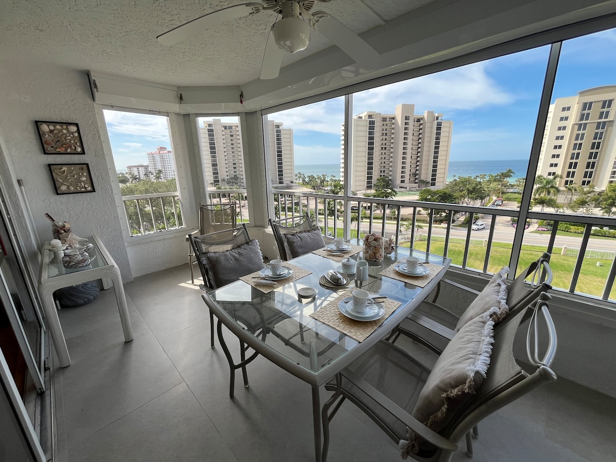 A sunlit dining area is shown with a glass table surrounded by six chairs, decorated with neutral-toned tableware. Large windows provide an expansive view of the surroundings, with buildings and the ocean visible in the distance, creating a sense of spaciousness and light.