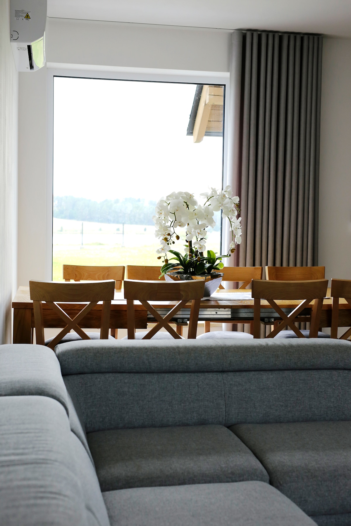 A dining area is visible with a wooden table surrounded by chairs, complemented by a vase of white orchids. Large windows allow natural light to fill the space, offering views of the surrounding landscape. A soft gray couch is situated in the foreground.