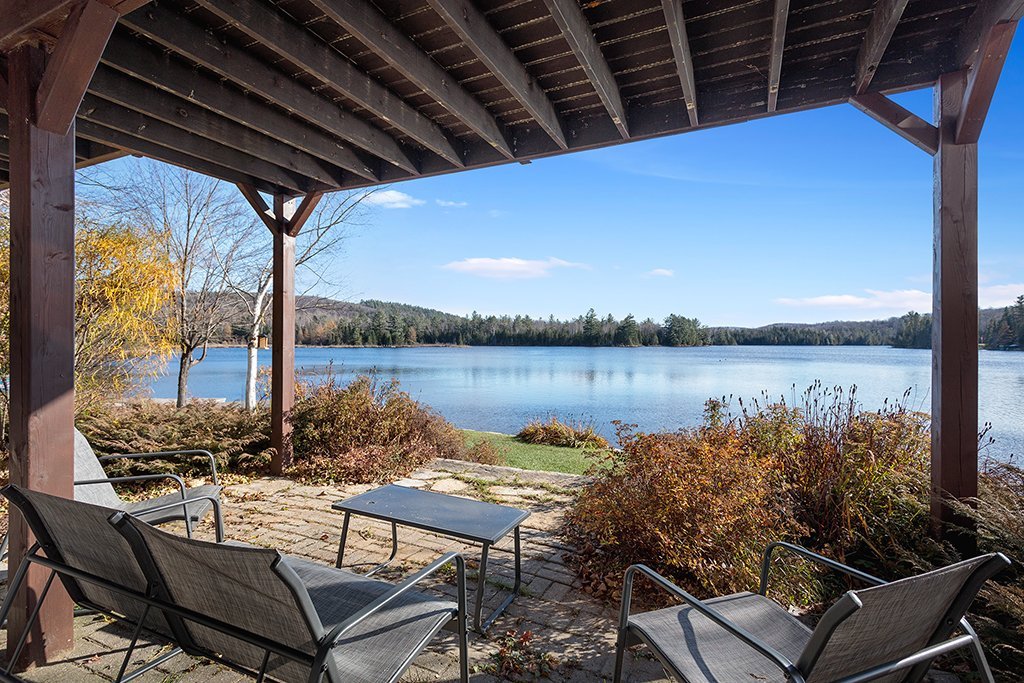 A covered patio area is featured, with two metal chairs and a small table arranged on a stone surface. A peaceful lake view is seen in the background, framed by trees and a clear blue sky.