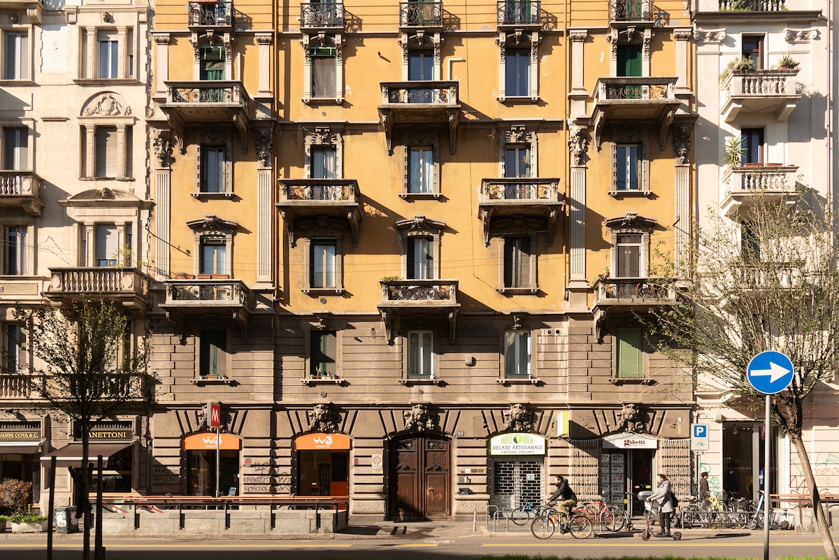 The image depicts a vibrant building facade featuring architecture in warm yellow tones. Multiple balconies with decorative wrought iron railings line the structure. Below, various storefronts are visible, with a clear street sign indicating direction and bicycles parked along the curb.