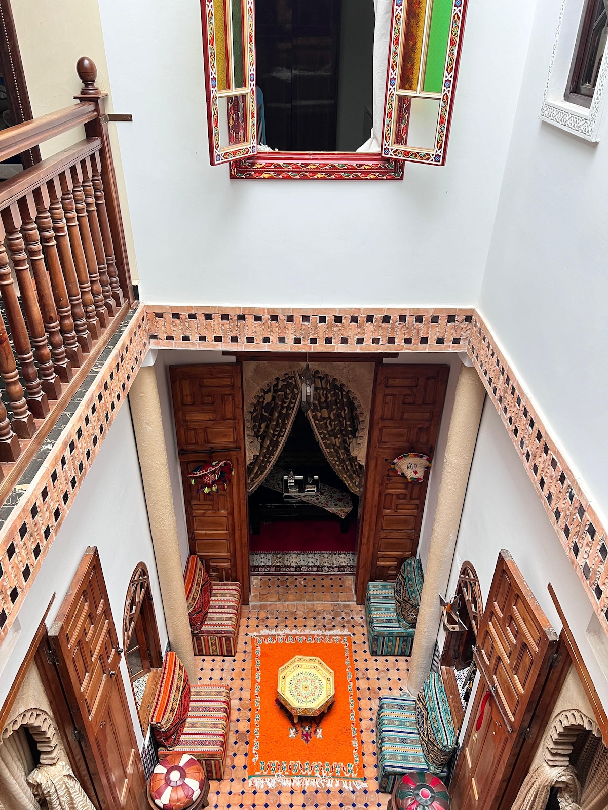 A courtyard features ornate wooden doors and colorful patterned seating arranged around a central table. The view highlights a vibrant orange rug, while intricate tile work decorates the walls. An upper balcony is visible, along with a decorative mirror reflecting the ambience.