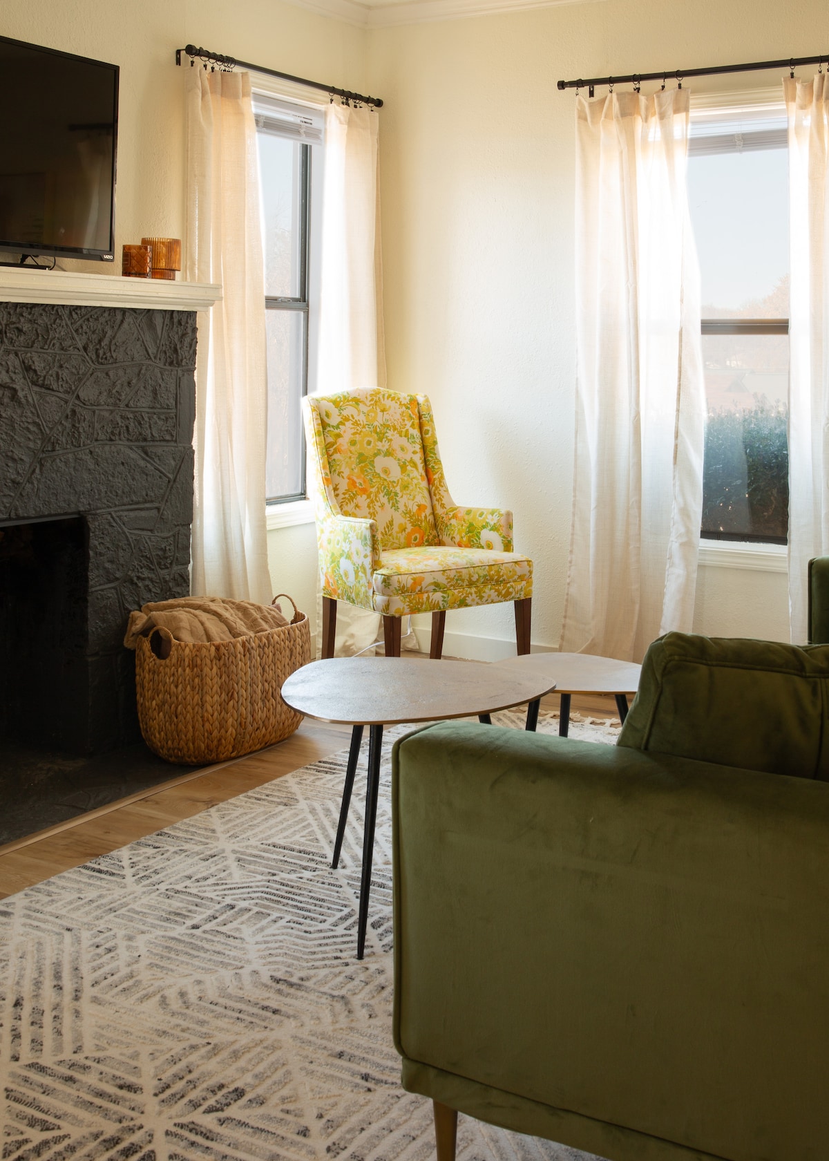 A living room features large windows dressed with sheer curtains, allowing natural light to fill the space. A colorful patterned chair adds a pop of brightness next to a low coffee table. A wicker basket rests on the floor, complementing the textured area rug.