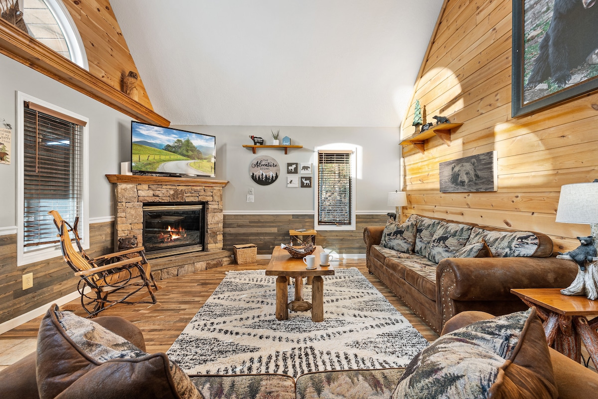 A spacious living area is highlighted by natural wood paneling and stone accents. Comfortable leather sofas are arranged around a cozy fireplace, with a large flat-screen television mounted above. Soft light fills the room from large windows, enhancing the inviting atmosphere.