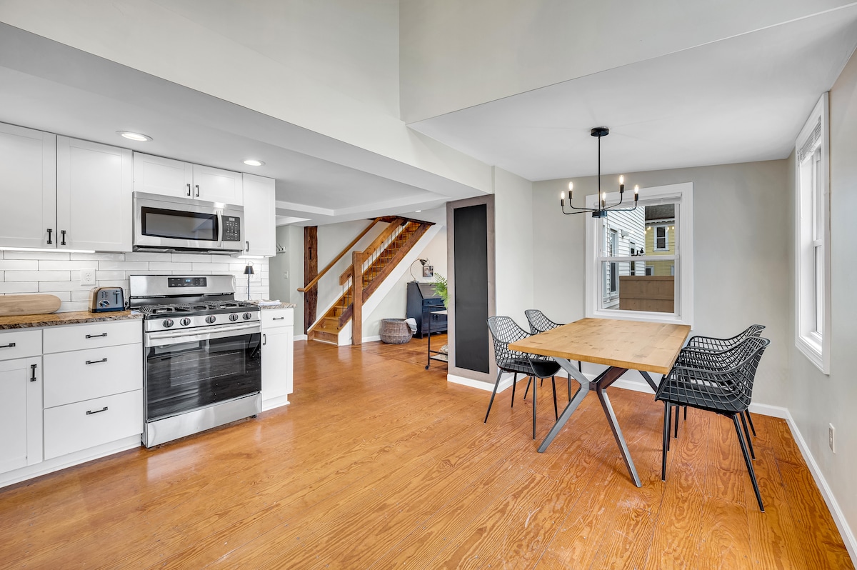 An open-concept kitchen and dining area features modern appliances, including a sleek gas stove and stainless steel microwave. A wooden dining table with black wire chairs is positioned beside a large window, allowing ample natural light. The spacious layout is accentuated by warm wooden flooring.