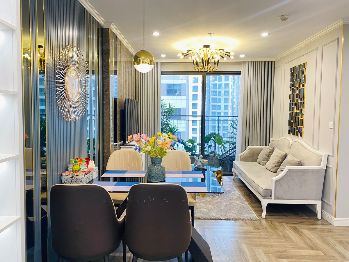 A modern living area includes a stylish dining table with two chairs and a comfy sofa, enhanced by decorative pillows. A wall-mounted television is visible, and large windows provide a view of the outside, surrounded by light-colored curtains. A vase of flowers adds a touch of color.