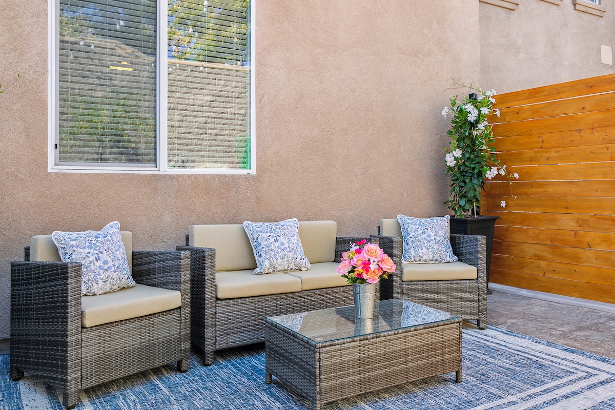 An outdoor seating area is presented with a glass-top table and a plush couch set featuring light-colored cushions, adorned with decorative pillows. A wooden privacy fence and a blooming plant add natural elements, while sunlight filters through the nearby window.