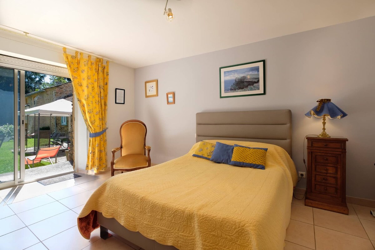 A well-lit bedroom features a spacious bed dressed in a yellow quilt, complemented by decorative pillows. A comfortable armchair and a wooden nightstand with a lamp are present. Large glass doors open to a grassy outdoor space, bordered by inviting curtains.