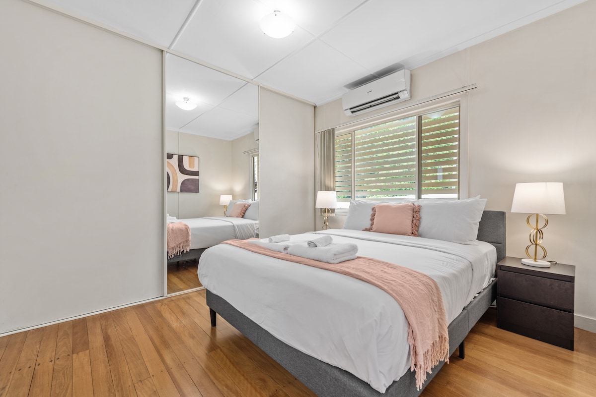 A spacious bedroom is presented with a queen bed dressed in hotel-quality linen, complemented by an accent throw. A mirror wardrobe reflects natural light from the window, enhancing the warm wood floor. Two bedside lamps provide soft lighting, creating a calming ambiance.
