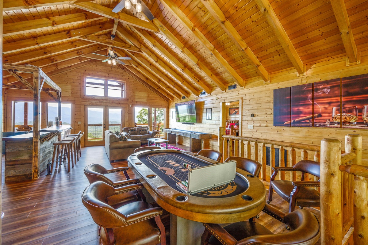 An open-concept living space features high wooden ceilings with exposed beams. A large table with leather chairs is positioned in the center, surrounded by seating areas that offer scenic views through expansive windows. A bar area is seen in the background, enhancing the entertainment space.