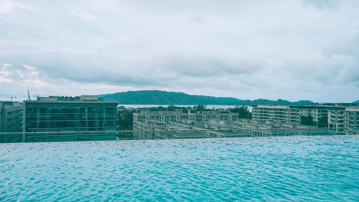 An expansive view is captured from the edge of an infinity pool, with a clear blue water surface extending towards the horizon. Low-rise buildings are visible in the foreground, while a distant mountain range and calm waters of the sea complete the serene landscape.