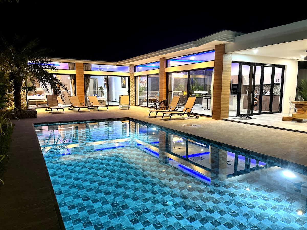 A private pool reflects shimmering blue tiles under soft outdoor lighting. Lounge chairs are arranged around the pool, with contemporary architecture visible in the background through large glass doors.