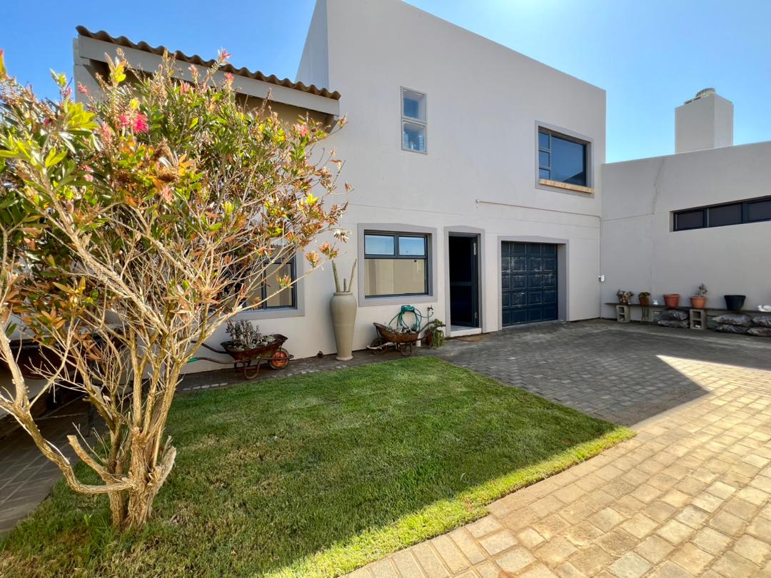The exterior of a well-kept property is presented, featuring a neatly trimmed lawn and a paved driveway. A flowering bush and various potted plants add greenery to the scene. A garage door and windows provide functional elements, all under a clear blue sky.