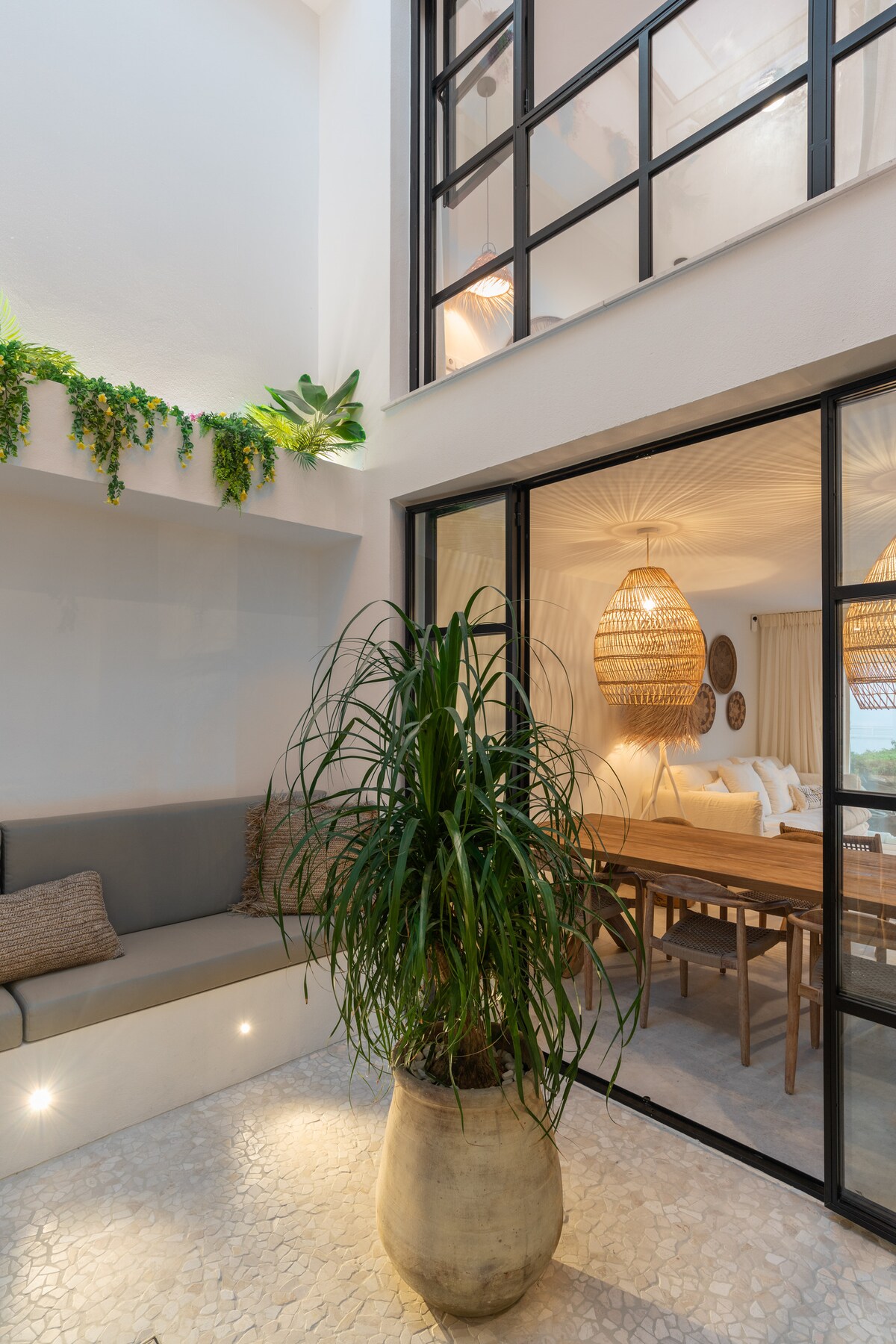 A spacious indoor seating area is highlighted by a large potted plant, offering a touch of greenery. Natural light filters through expansive glass doors, revealing a dining space with woven pendant lights and a wooden table, enhancing the inviting atmosphere of the home.