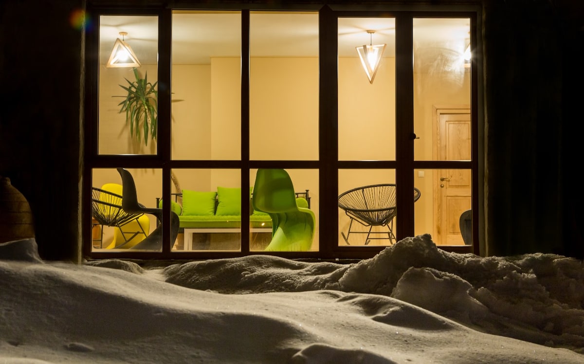 A cozy lounge area is visible through large windows, showcasing modern chairs and a bright green sofa. Two pendant lights enhance the space's warmth, while soft snow can be seen covering the ground outside, creating a peaceful winter scene.