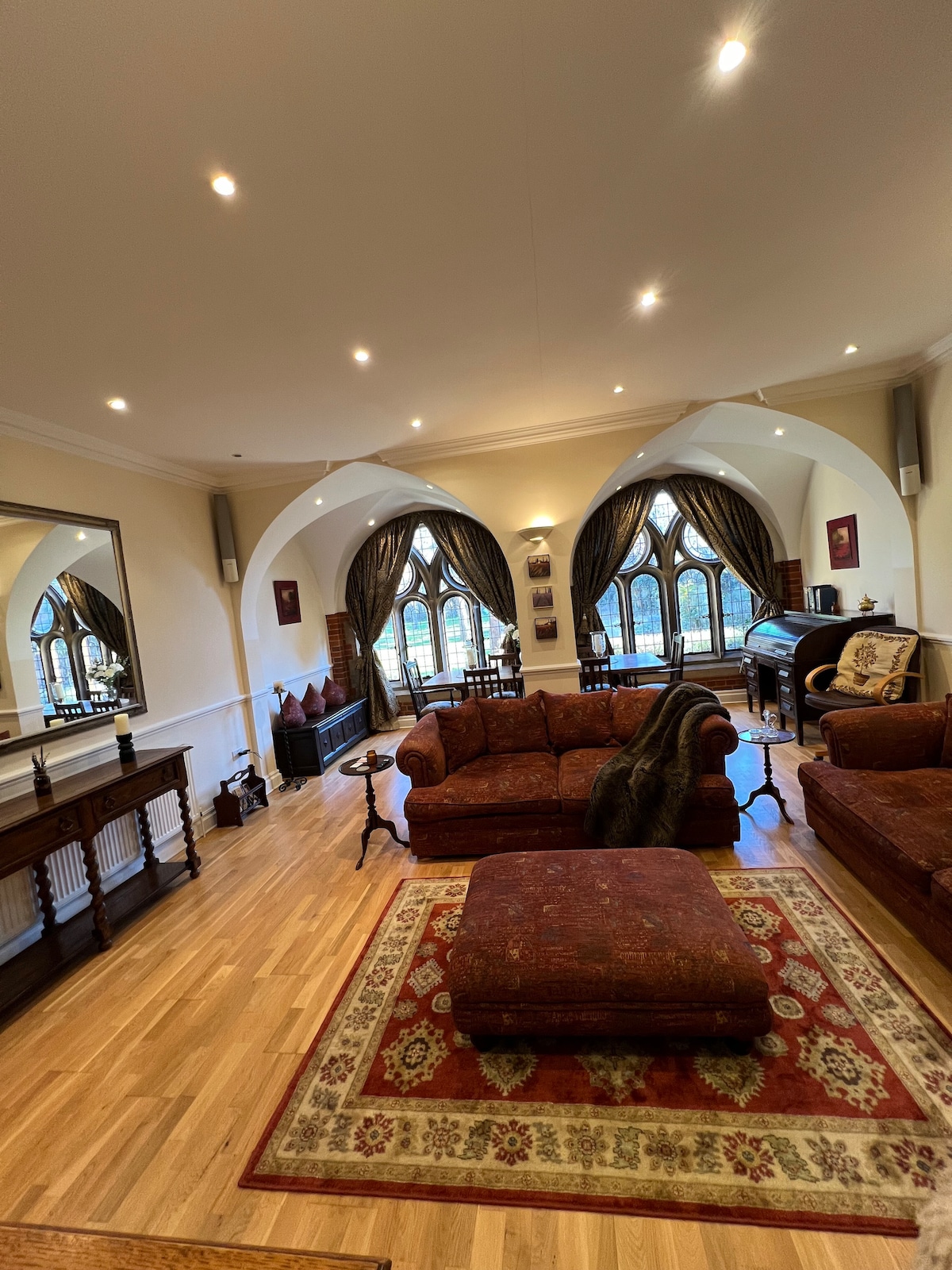 A spacious living area features arched windows that allow light to fill the room. Traditional furniture, including a large sectional sofa and a coffee table, is arranged around a decorative rug. A piano and seating area are visible, contributing to the elegant ambiance.