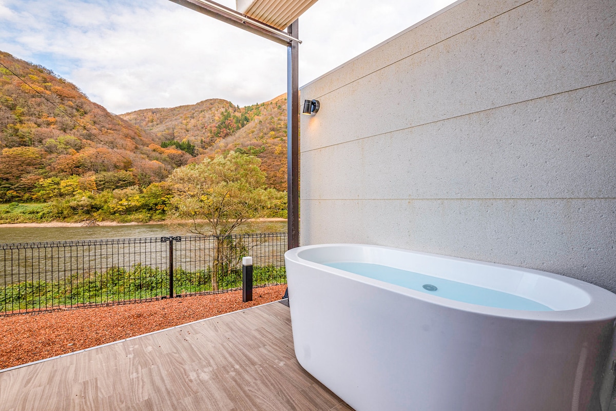 A modern outdoor bathtub is positioned on a wooden deck, overlooking the scenic Mogami River. The view features gently sloping hills adorned with colorful autumn foliage, providing a tranquil backdrop. Soft ambient lighting is visible near the tub, enhancing the serene environment.