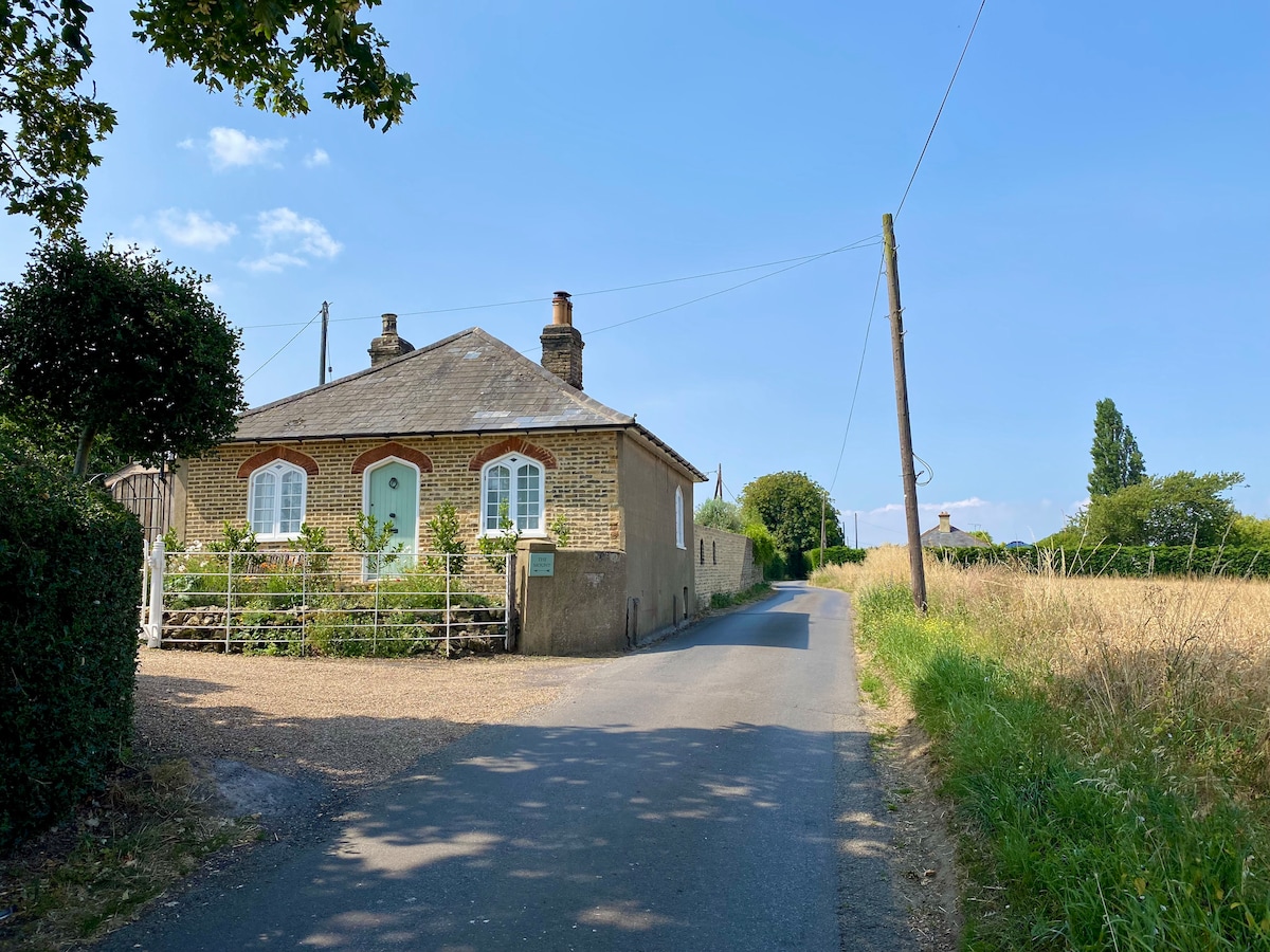 A charming Victorian lodge is positioned along a quiet single track road, featuring a welcoming entrance with a white gate and green doors. Surrounding fields and trees are visible in the background under a clear blue sky.