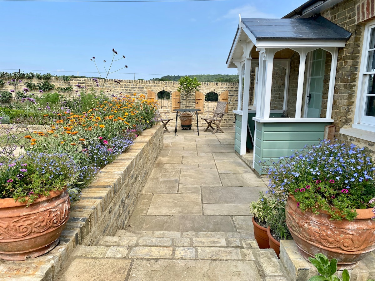 A sunlit stone terrace leads to a charming seating area with two wooden chairs and a small table. Flower pots brim with vibrant blooms, while the backdrop showcases the well-maintained garden, characterized by colorful flowers and a low brick wall.