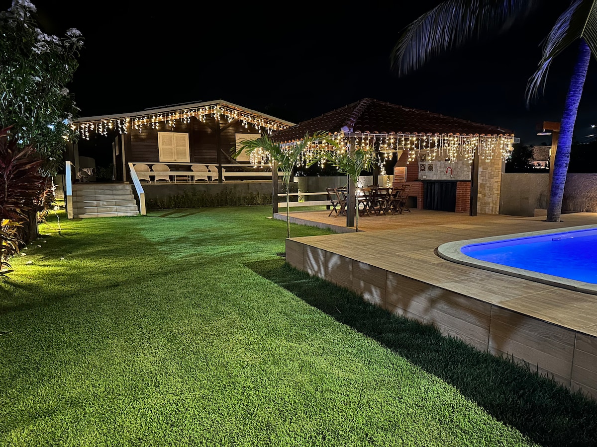 The outdoor space features well-maintained grass, a swimming pool with illuminated water, and a wooden deck where an outdoor seating area is present. String lights hang above, adding a soft glow to the setting, complemented by tropical plants and a rustic house in the background.