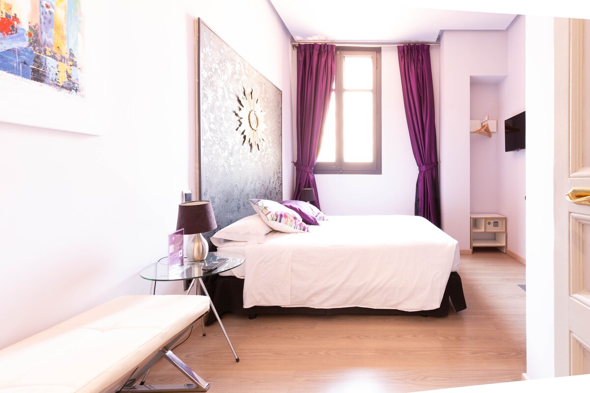 A well-lit guest room features a spacious bed against a decorative wall with intricate artwork. Light purple curtains frame a window, allowing natural light to fill the space. A small glass table and a minimalist bench are present, complementing the modern decor.