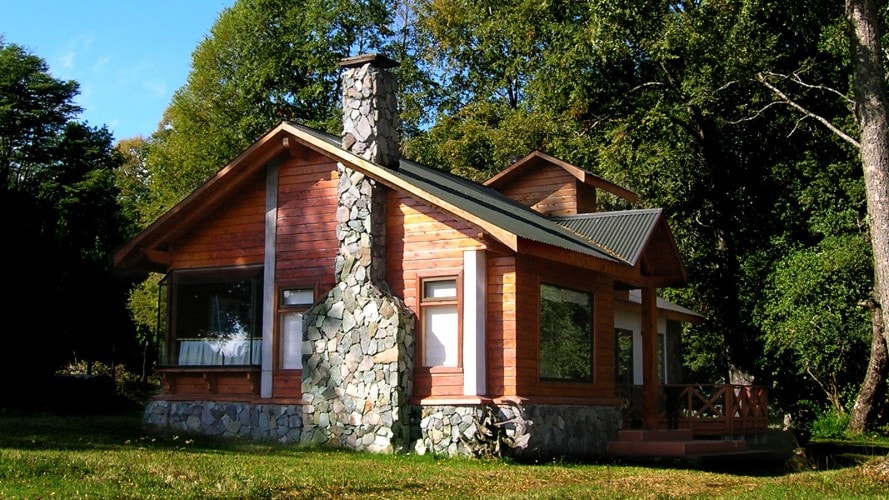 A charming cabin with a wooden exterior and stone chimney is situated among lush greenery. Large windows allow natural light to fill the interior, while a deck extends from the entrance, providing a welcoming outdoor space.