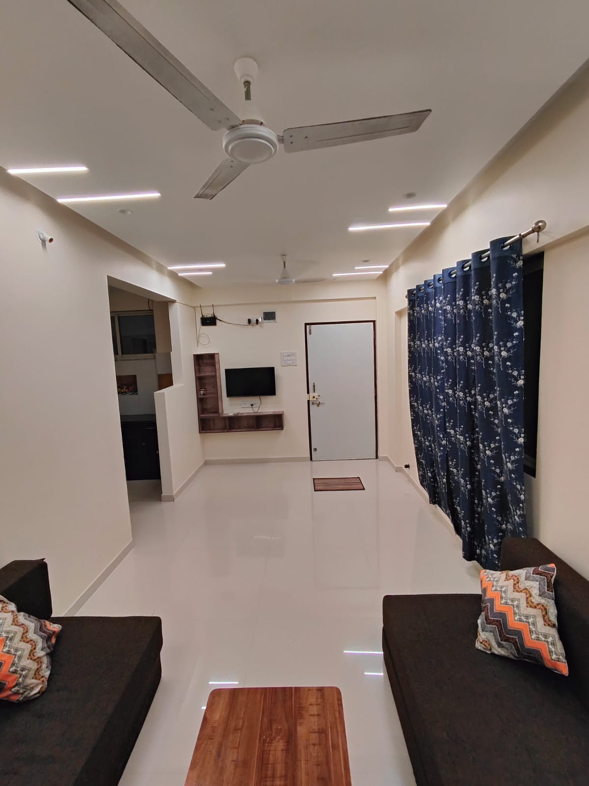 A clean, well-lit living area is visible with two dark sofas adorned with patterned pillows. Large windows are draped with dark blue curtains, while a flat-screen TV is mounted on a wall to the left, creating a functional and inviting space.