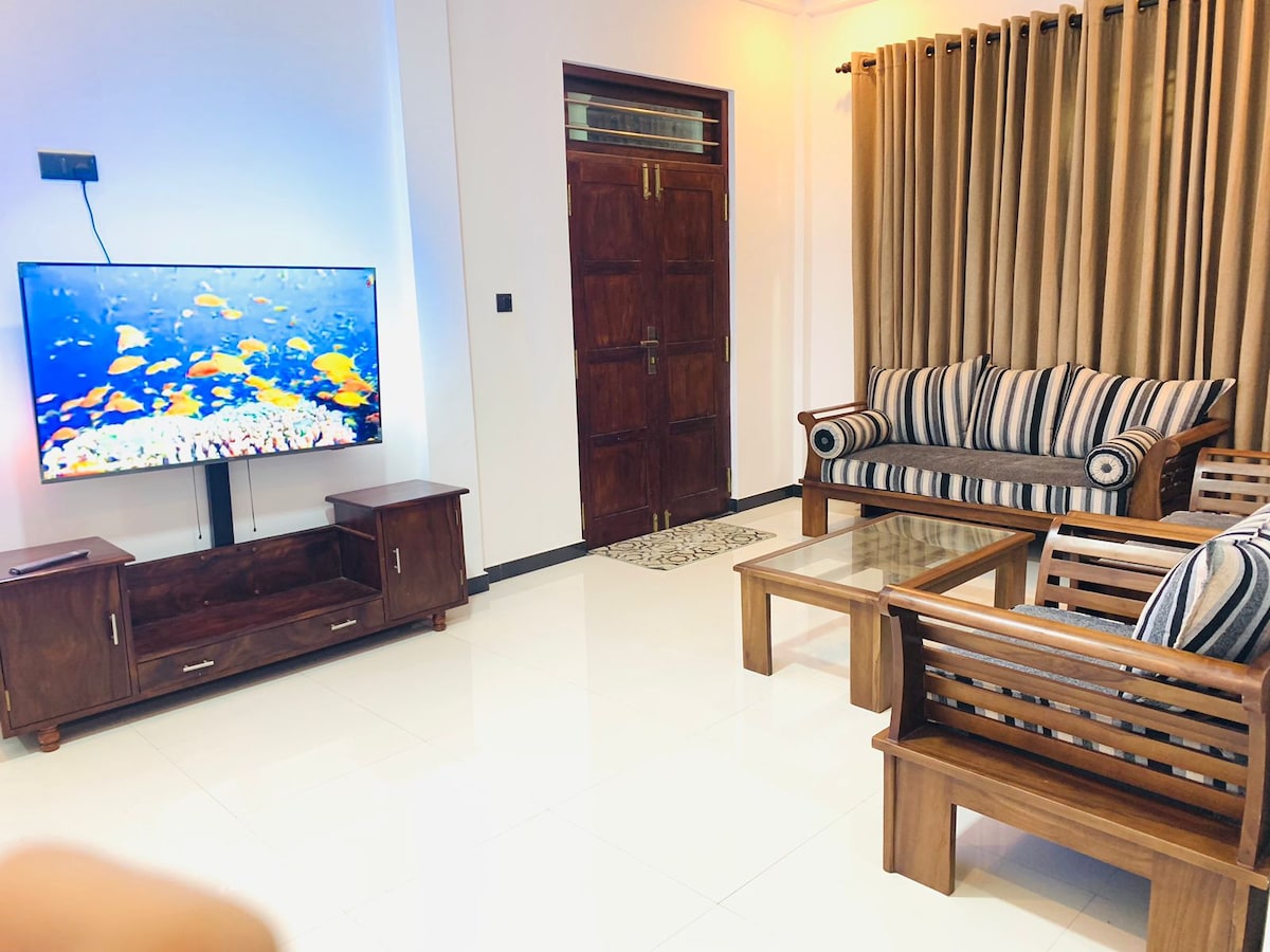 A living area is presented with a flat-screen television mounted on the wall. Comfortable seating includes a striped sofa and two wooden chairs. A glass coffee table sits in the center, while large curtains frame the windows, creating a cozy environment.