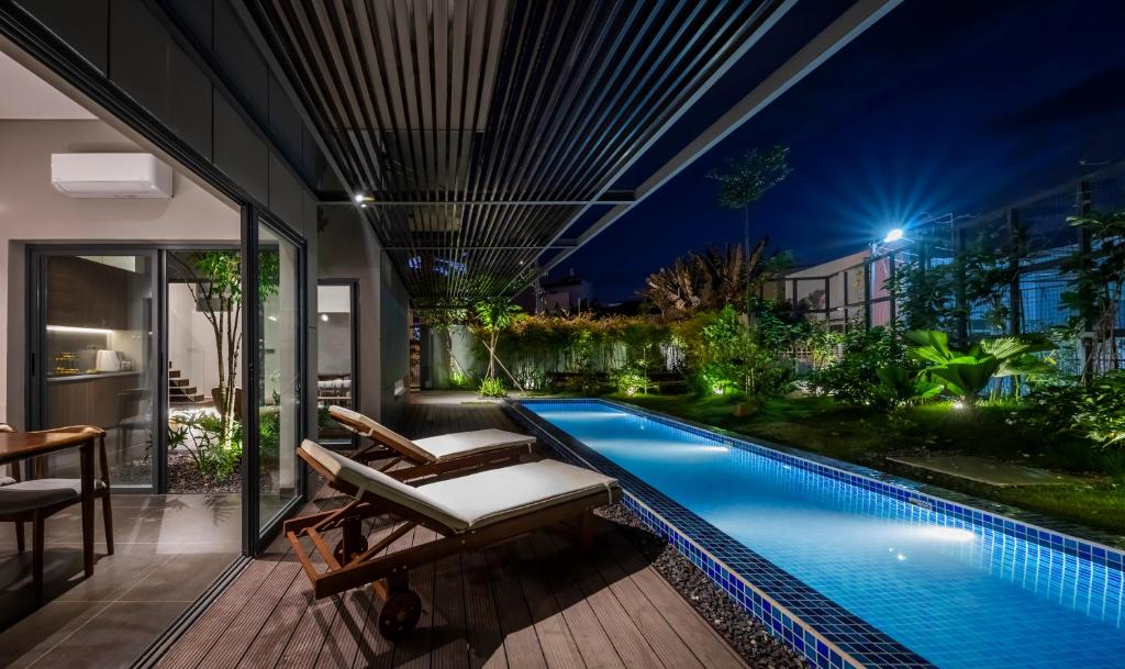 An outdoor area features a long swimming pool surrounded by lush greenery, illuminated softly by outdoor lighting. Two wooden lounge chairs are positioned alongside the pool, providing a relaxing spot to enjoy the serene atmosphere. The space is designed for comfort and tranquility.