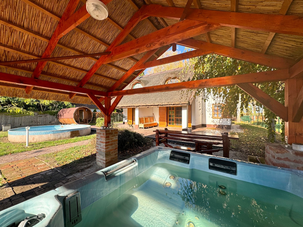 A covered area features a bubbling hot tub beneath a thatched roof, providing a relaxing space. An outdoor seating area with wooden benches is visible nearby, while a pool and a wooden structure are seen in the background, surrounded by greenery.