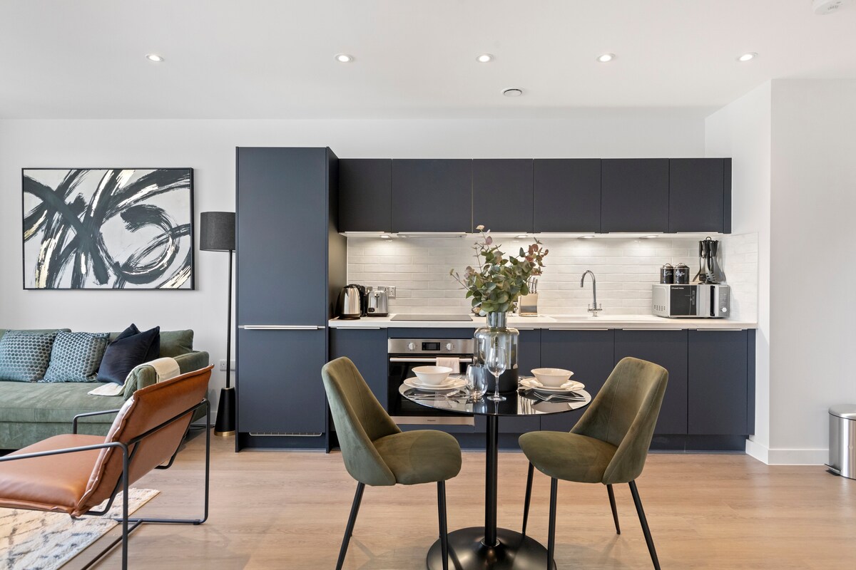 The modern kitchen features sleek dark cabinetry and a stylish dining area. A round table is set with elegant dishware, surrounded by two green chairs. A cozy green sofa is positioned nearby, complemented by decorative pillows and a vibrant artwork above the furniture.