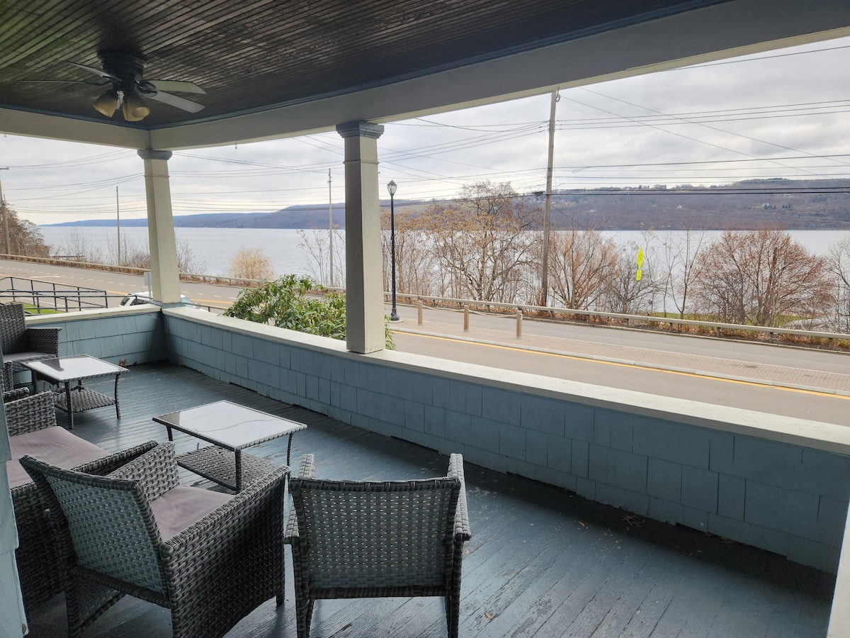 A spacious porch is furnished with comfortable wicker seating and a table, providing a relaxing area to enjoy views of the harbor and Seneca Lake. The scenery includes gently sloping hills and trees overlooking the water.