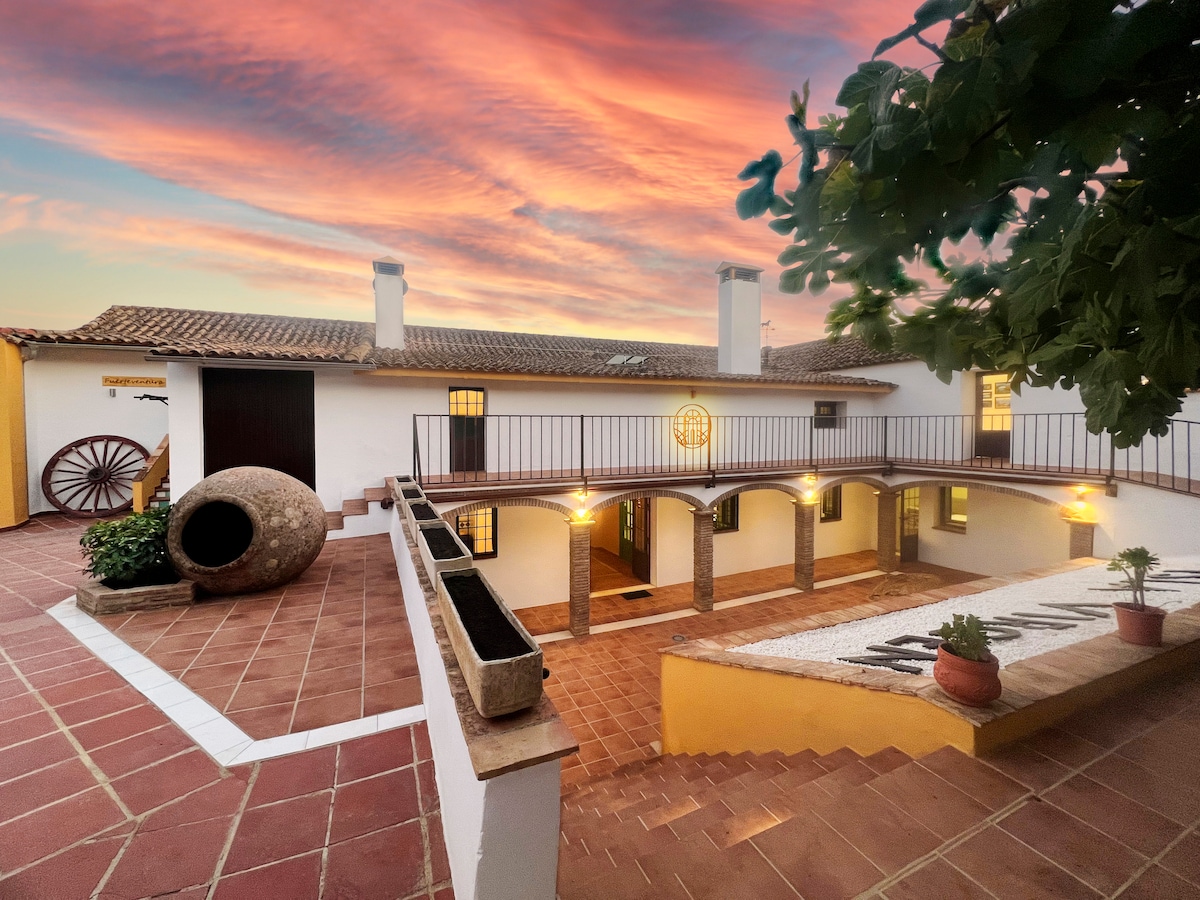 A charming courtyard is framed by a two-story building, featuring arched columns and warm lighting. A large decorative pot and potted plants are placed along the tiled walkway. The sky is painted with soft hues of orange and pink during sunset.