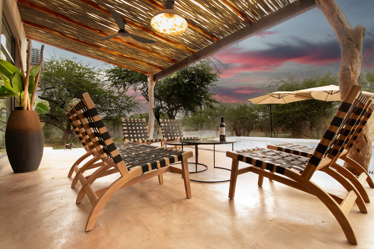 An outdoor seating area features two wooden lounge chairs with patterned fabric and a small table, set under a covered patio. A tranquil pool and lush vegetation are visible in the background, with vibrant sunset hues illuminating the sky.