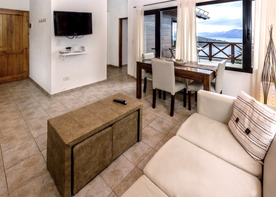 An open living area features a comfortable light-colored sofa adjacent to a wooden dining table, which seats four. A large window offers views of the surrounding landscape. A flat-screen TV is mounted on the wall, and tile flooring adds to the spacious feel.